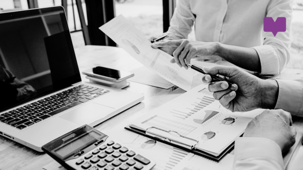 Close up of two people's arms working at a desk with a laptop. They are holding papers with various charts on, and have a calculator to hand.